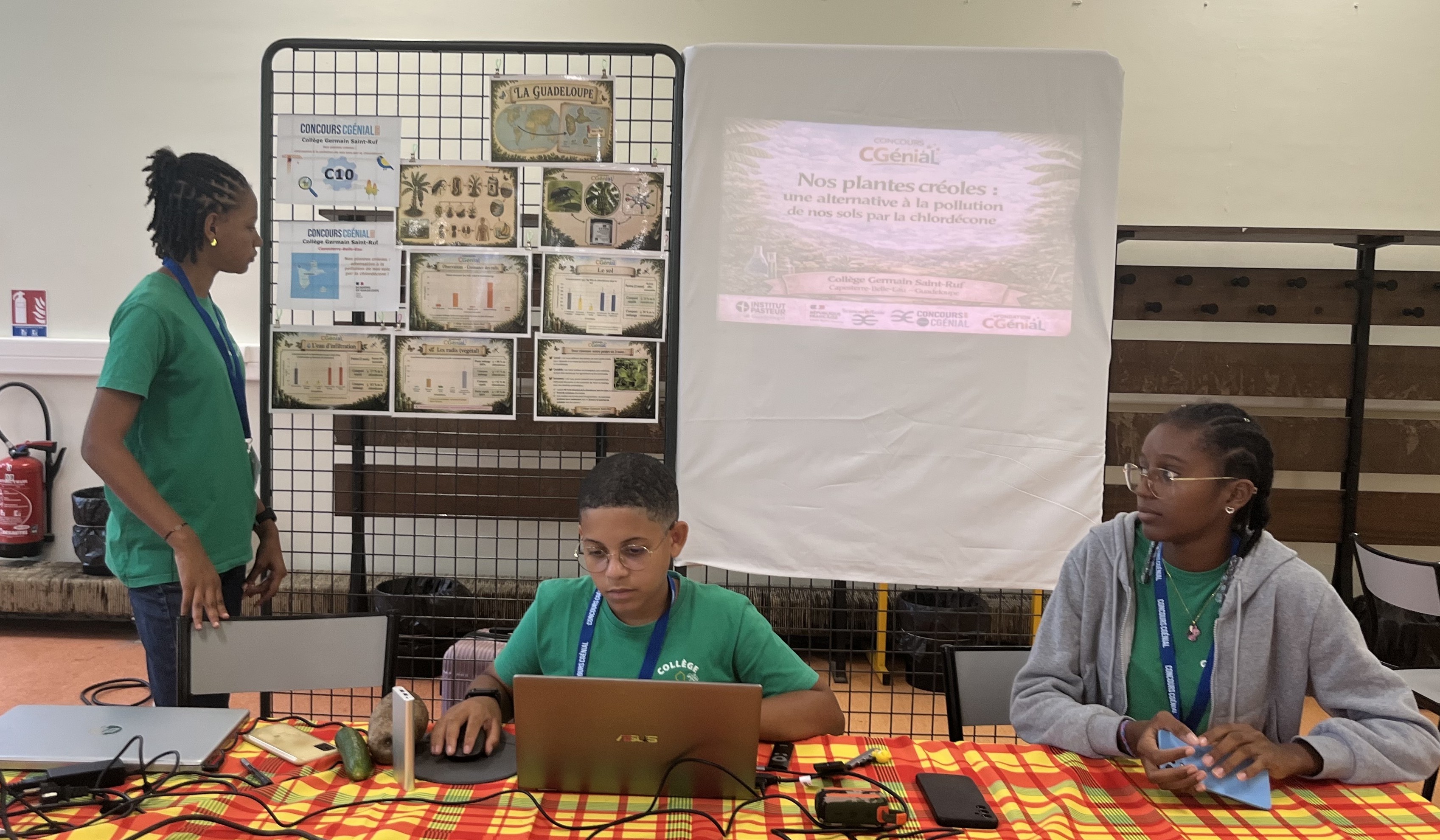 Collège Germain Saint-Ruf - CCG2026 Trois collégiens présentent un projet scientifique lors du concours CGénial. Ils se tiennent autour d'une table recouverte d'une nappe madras jaune et rouge, sur laquelle sont disposés un ordinateur portable, des échantillons de plantes et des flacons. En arrière-plan, un panneau d'affichage et un écran de projection affichent le titre 'Nos plantes créoles : une alternative à la pollution de nos sols par la chlordecone'."