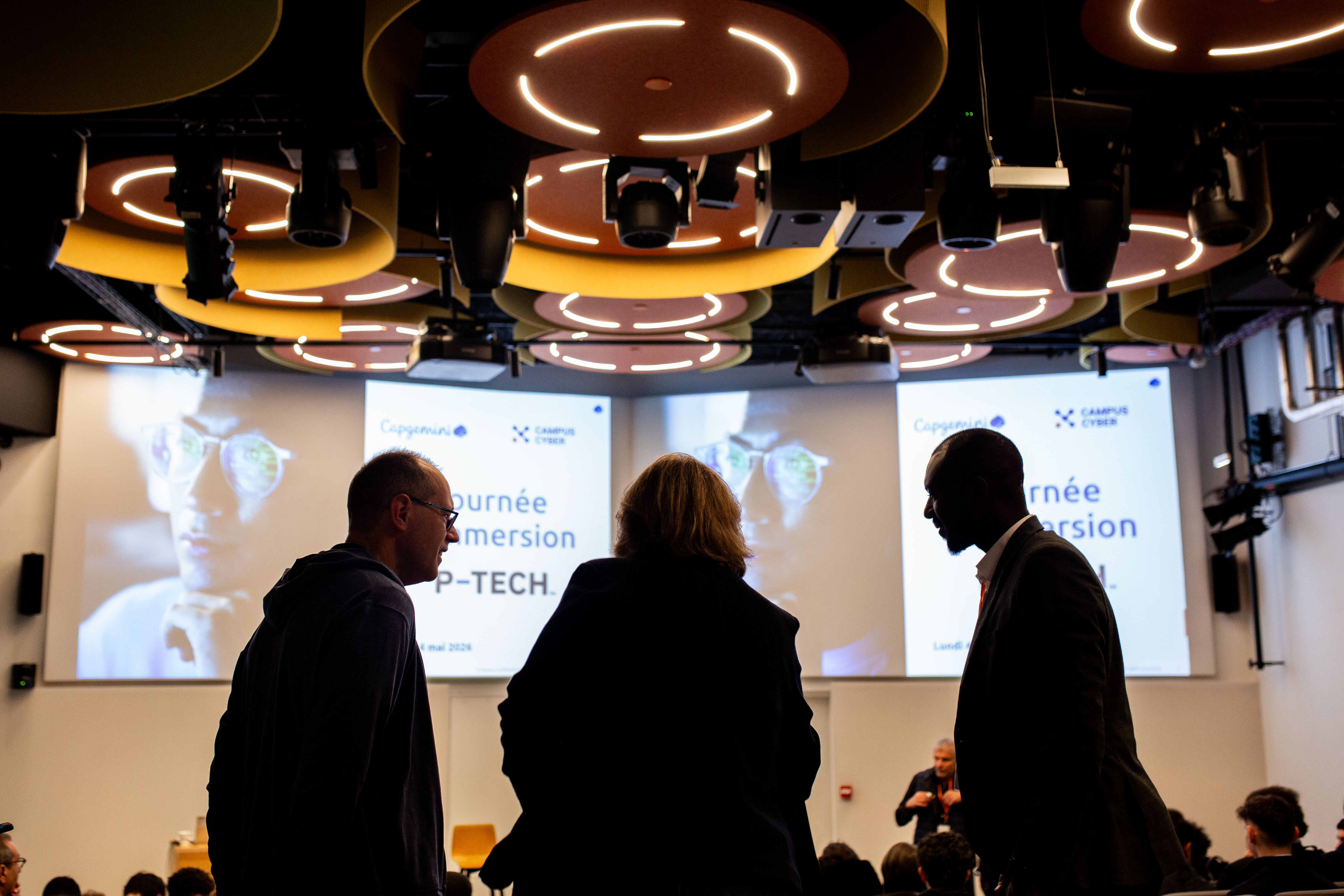 Trois personnes discutent en silhouette au premier plan d'une salle de conférence. En arrière-plan, un grand écran affiche 'Journée d'Immersion P-TECH' sous les logos de Capgemini et du Campus Cyber.